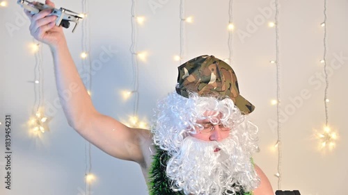 A man wearing a Santa wig and fake beard, camouflage cap, and topless, holding a drone and its remote controller above his head, pretending the drone is flying like in a video game