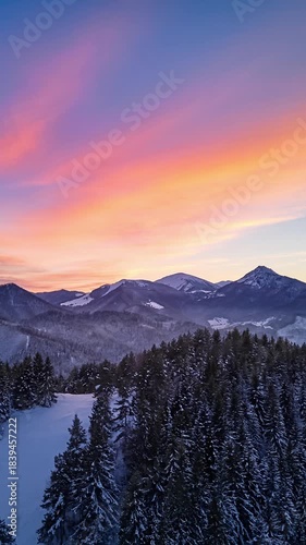 Vertical Aerial hyperlapse over winter mountain forest and snowy peaks under colorful evening sunset sky