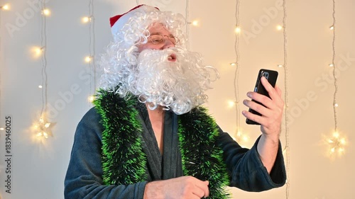 A man dressed as Santa Claus on a white background with festive lights, pleasantly talking on a smartphone during a video call, creating a cheerful and warm holiday scene.