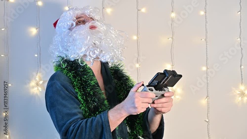 A man dressed as Santa Claus on a white background with festive lights, holding a drone remote controller and looking upward, creating a playful and modern holiday scene.