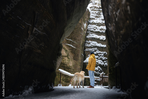 Man walks with dog along narrow snowy path between high rocks. Labrador retriever stays close to owner in cold winter landscape.