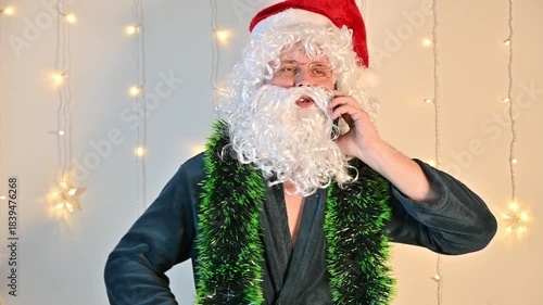 A man dressed as Santa Claus on a white background with festive lights, talking on a smartphone with a tense and stressed expression, creating a humorous and dramatic holiday scene.
