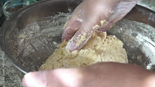 Hands are mixing and kneading dough with flour in a kitchen. The process takes place during the day, showing the raw ingredients being combined.