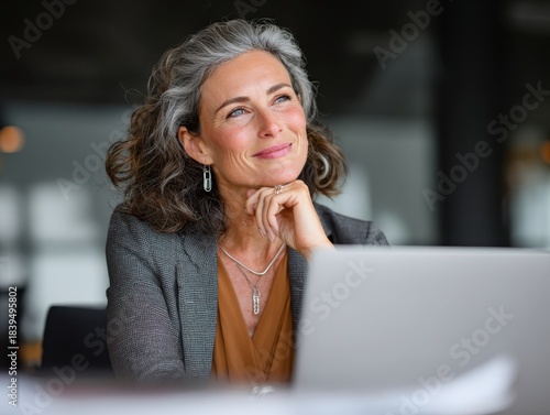 Businesswoman with Silver Hair Sitting in Office with Laptop Smiling Mature Woman with Positive Attitude Looking Away Contemplating Business Ideas While Working in Modern Workplace