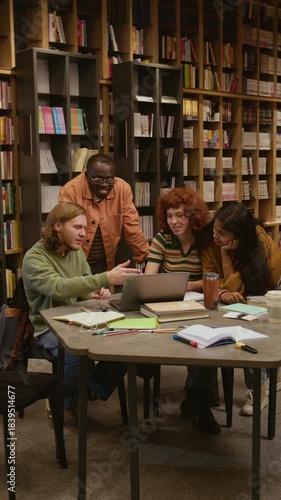Vertical shot of smiling multicultural college students watching video on laptop while collaborating on project after classes in library