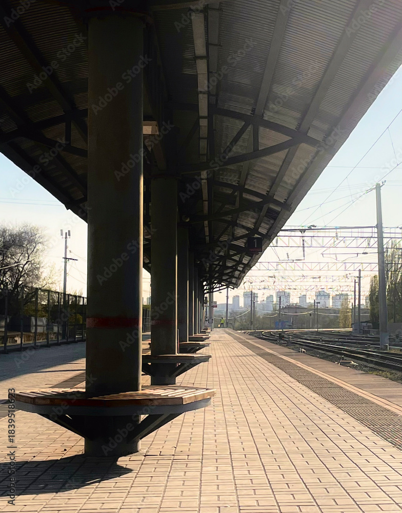 Naklejka premium Quiet train station platform with waiting area and tracks visible under bright sunlight