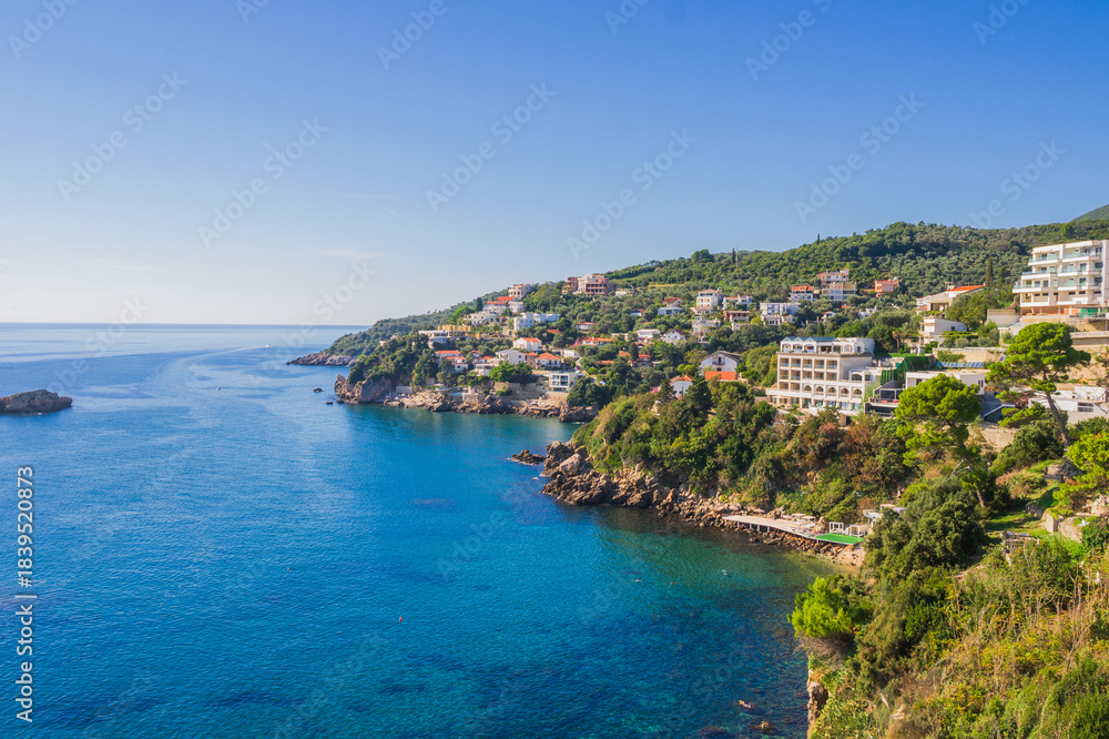 Fototapeta premium Coastal view of Ulcinj, Montenegro, with hillside houses and resorts overlooking the Adriatic Sea. Crystal-clear blue waters meet lush greenery under a bright summer sky.