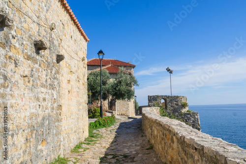  Narrow stone walkway in Ulcinj’s Old Town, Montenegro, leading past ancient walls to a seaside lookout. The Adriatic Sea glistens under a clear blue sky beside historic fortress structures.