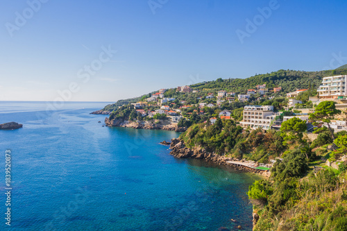  Coastal view of Ulcinj, Montenegro, with hillside houses and resorts overlooking the Adriatic Sea. Crystal-clear blue waters meet lush greenery under a bright summer sky.