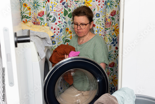 Woman stands in bathroom putting clothes into washing machine while surrounded by towels and laundry, carrying out daily household task with focus and care