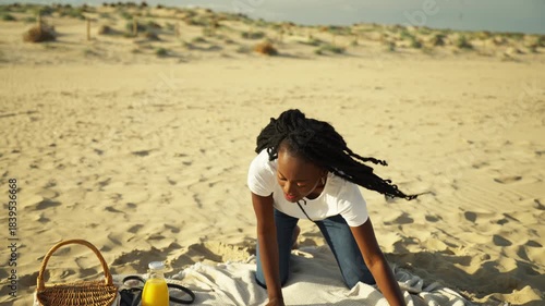 Beautiful african american woman with long braids setting up blanket for picnic on sandy beach, enjoy warm sunlight and smiling while holding an orange in hand