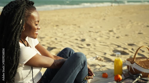 Gorgeous young african american woman with flowing dreadlocks lounging and grinning on sandy beach while having picnic. Tranquil lady savoring calm summer day and holiday at ocean