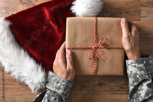 Closeup of a soldier holding a Christmas present with a Merry Christmas tag on a wood table and a Santa hat.
