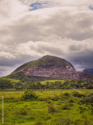 Mountain with exposed rock layers and lush green fields under dramatic cloudy sky in a rural landscape.