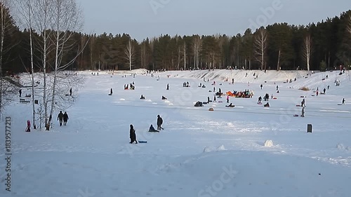 A wide shot of snow-covered hill, where many people are sledding and enjoying winter fun, families and individuals glide down the slope against backdrop of bare trees and forest
