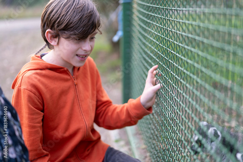 A boy in an orange hoodie shares a delightful moment at the zoo, leaning closely to a fence to connect with an animal. His excitement is palpable as he explores the wonders of nature alongside family.