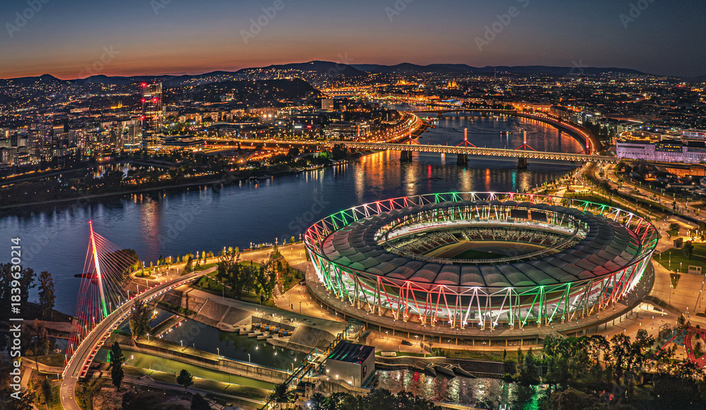Obraz premium Budapest. Drone night view of a stadium and riverfront cityscape illuminated with colorful lights, showing bridges, buildings and vibrant urban life from above.