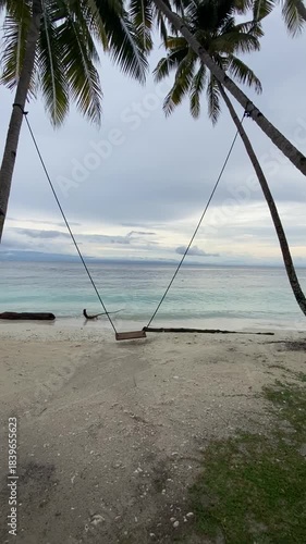 wooden swing tied between two tall palm trees on a white sandy beach, gently swaying back and forth near the turquoise ocean.