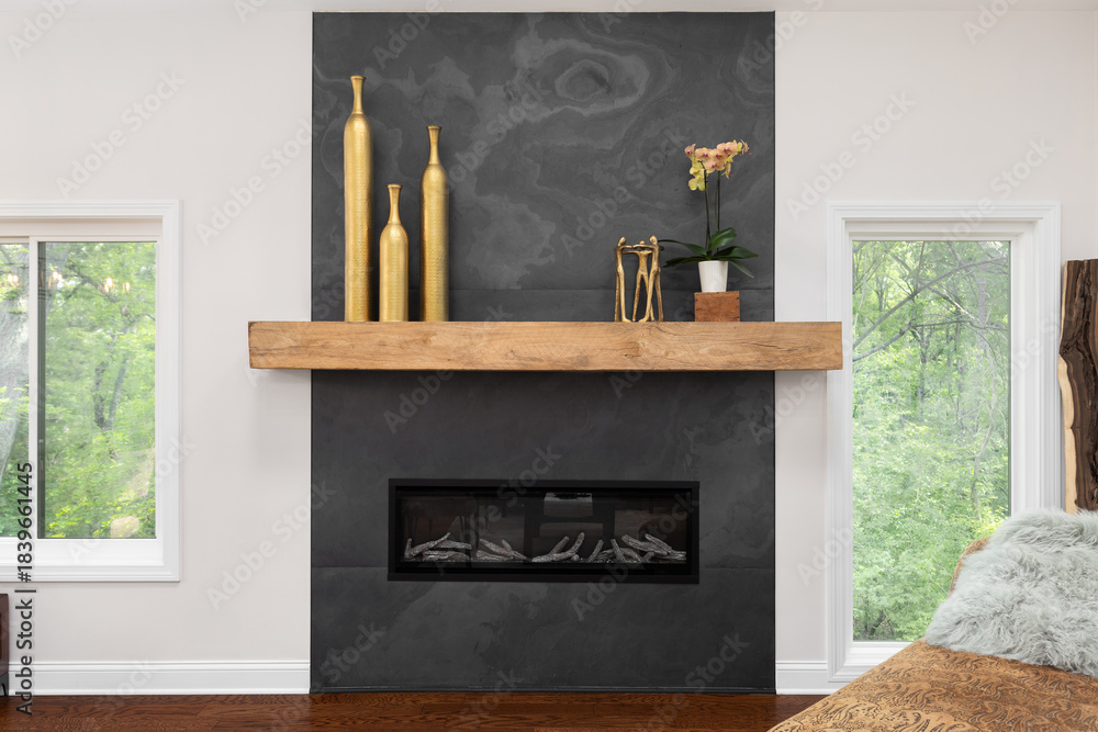 Obraz premium A modern fireplace with a dark slate surround and a natural wood mantel. Golden decorative vases and a plant add elegance with windows on the side.