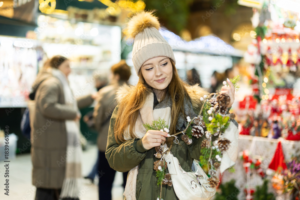Fototapeta premium Young woman walks through outdoor X mas bazaar, choose and examines garland of fir cones near shop window of New Years goods. New Year open air fair fest