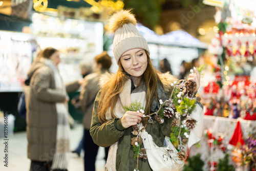 Young woman walks through outdoor X mas bazaar, choose and examines garland of fir cones near shop window of New Years goods. New Year open air fair fest