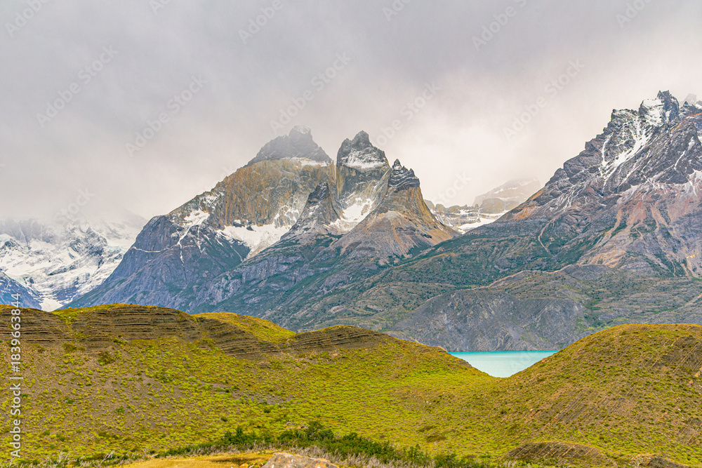 Fototapeta premium Dramatic mountain peaks rising above rolling terrain with a glimpse of turquoise water in Torres del Paine National Park, Patagonia, Chile.