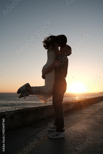Man lifting woman in joyful embrace by the sea at sunset, romantic silhouette moment filled with love and happiness.