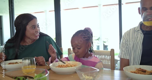 Following mother raising juice glass, diverse family sipping twirling pasta at marble breakfast bar