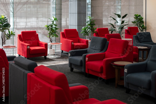 Modern lounge seating area with numbered red and dark gray armchairs, side tables, and indoor plants, illuminated by natural light through large glass windows