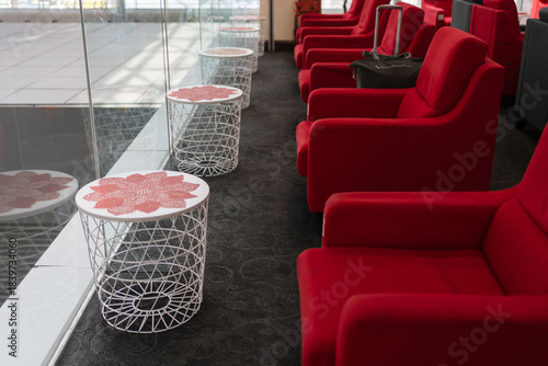 Modern lounge seating area with numbered red and dark gray armchairs, side tables, and indoor plants, illuminated by natural light through large glass windows