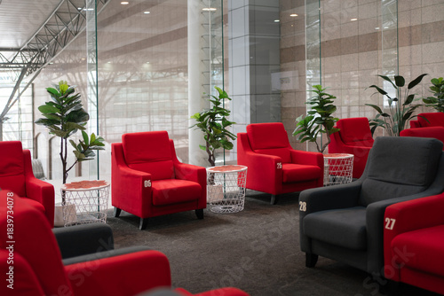 Modern lounge seating area with numbered red and dark gray armchairs, side tables, and indoor plants, illuminated by natural light through large glass windows