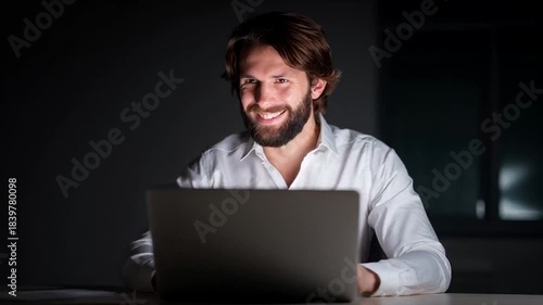 Smiling man engrossed in laptop, nocturnal work energy, modern male focused in dimly lit office, international men's day reflection