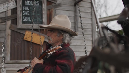 Man Wearing Cowboy Hat and Flannel Shirt Holding Mandolin String Instrument and Sitting on Porch Swing Outside of Old Rustic Saloon
