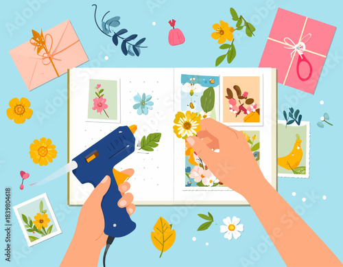 Hands using a glue gun to create a floral scrapbook with various craft supplies on a blue background.