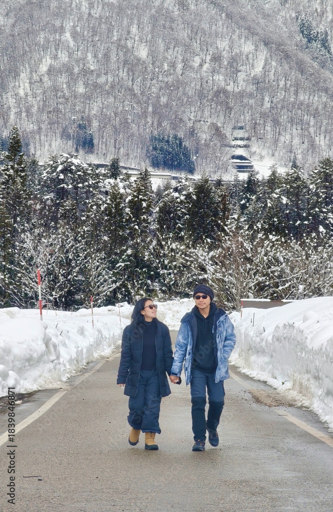 Fototapeta premium A couple walks hand-in-hand down a snow-covered mountain road, flanked by deep snowdrifts and a dense, snowy forest backdrop in winter