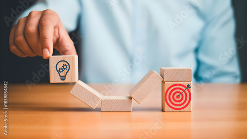 Conceptual image illustrating strategic business development from innovative ideas to successful goal achievement, showing a hand guiding a lightbulb block towards a target on a wooden path