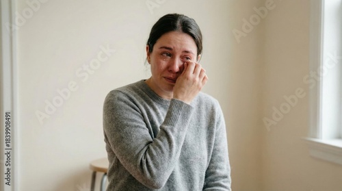 Emotional woman with long dark hair wearing a gray sweater, sitting indoors, wiping tears from her eyes, conveying feelings of sadness and vulnerability in a serene environment