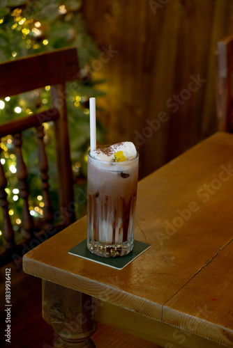 A refreshing iced chocolate drink topped with marshmallows , cocoa powder, and a small yellow flower, served with a straw on a wooden table in a cozy, festive setting.