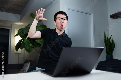 An Asian man wearing glasses makes a dramatic mic drop hand gesture while sitting at his desk with a laptop. He appears triumphant after finishing a task while working at night in his home office.