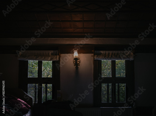 Tableau sur toile Soft lamplight glows between two wooden-framed windows in a historic Safranbolu mansion, highlighting rustic beams, textured walls, and the serene greenery visible through the dimly lit room