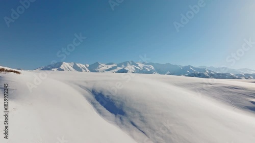 Aerial drone footage captures vast snow-covered landscape in Kyrgyzstan. Serene winter scene highlights majestic mountains under sunny sky. Boom up camera movement.