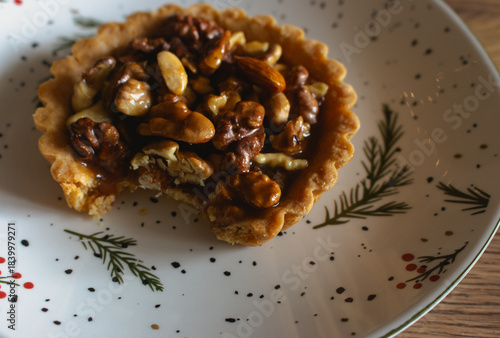 Traditional Christmas tart with nuts and salted caramel on decorated plate. Bitten tart on festive Christmas plate. Bitten hazelnut cake close up. Cinnamon cookie with caramel. Winter holiday food.
