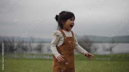 slow motion of happy toddler girl running and playing soap bubble in grass field near the river