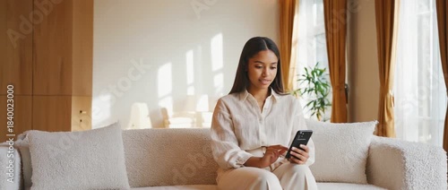 Modern Serenity: A woman in a bright room checks her phone sitting comfortably on a soft couch, the room bathed in natural light, exuding an atmosphere of peace and modern living.