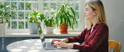 Working By The Window: A young woman working on laptop at the desk next to a window, surrounded by indoor plants, illuminated by natural daylight, a blend of productivity and serenity.