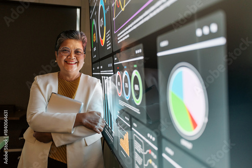 An Asian businesswoman stands confidently in a modern office, holding a tablet beside large digital dashboards with graphs and analytics, reflecting leadership, data-driven decision-making, planning.