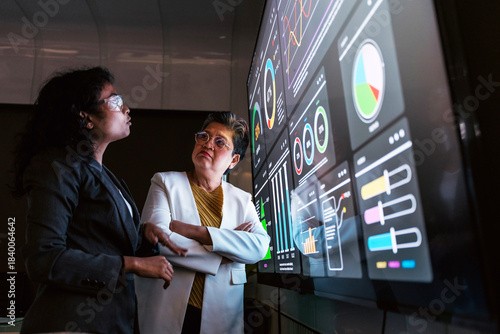 Two businesswomen—Indian and Asian—collaborate in a modern office, analyzing digital charts and dashboards on a large interactive screen, reviewing data insights for business planning and performance.
