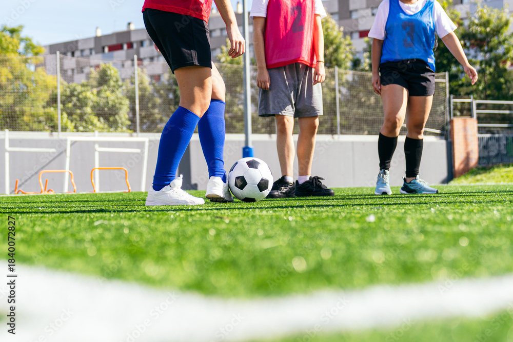 Obraz premium Three women footballers standing on green artificial turf with a soccer ball, preparing for practice on a sunny day