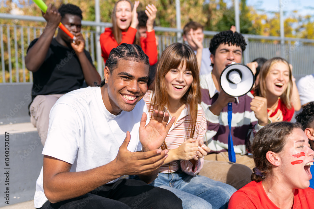 Obraz premium Diverse group of enthusiastic young adult friends watching a sports event in stadium stands, shouting and clapping with excitement