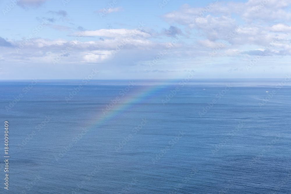 Fototapeta premium Gentle rainbow over the open ocean, blending with pale sunlight and drifting clouds in a peaceful atmosphere of calm water and vast horizon